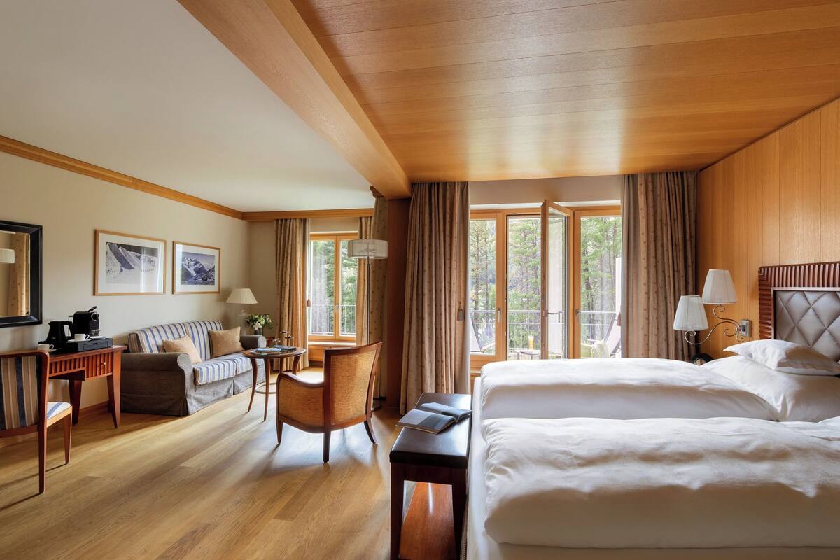 A hotel room with a large bed, a sofa, a desk, and a chair. There are windows with curtains, a lamp, and framed pictures on the wall.