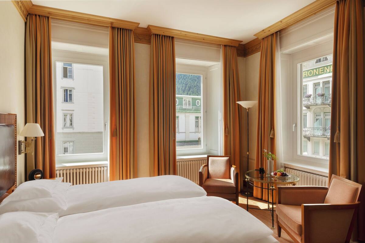 A hotel room with two beds, a sitting area with a table and chairs, and large windows with curtains.