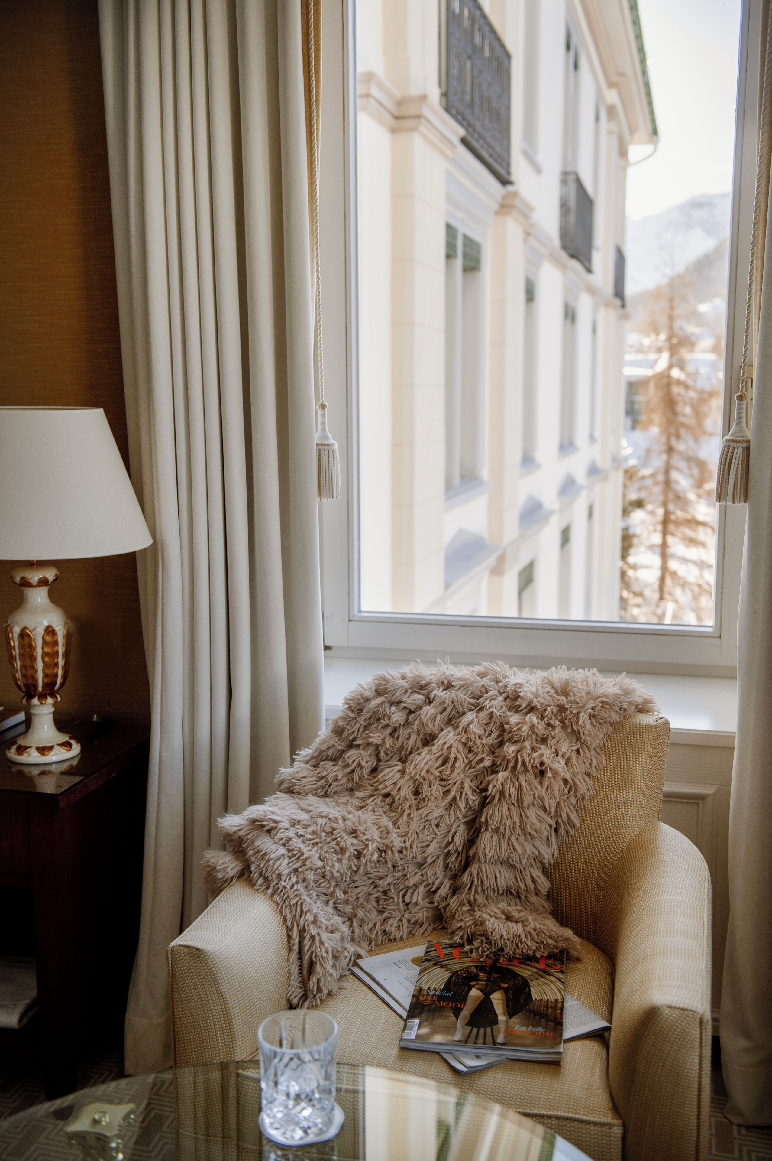 A cozy room with a window, a lamp, a fluffy throw, and a glass on a table.