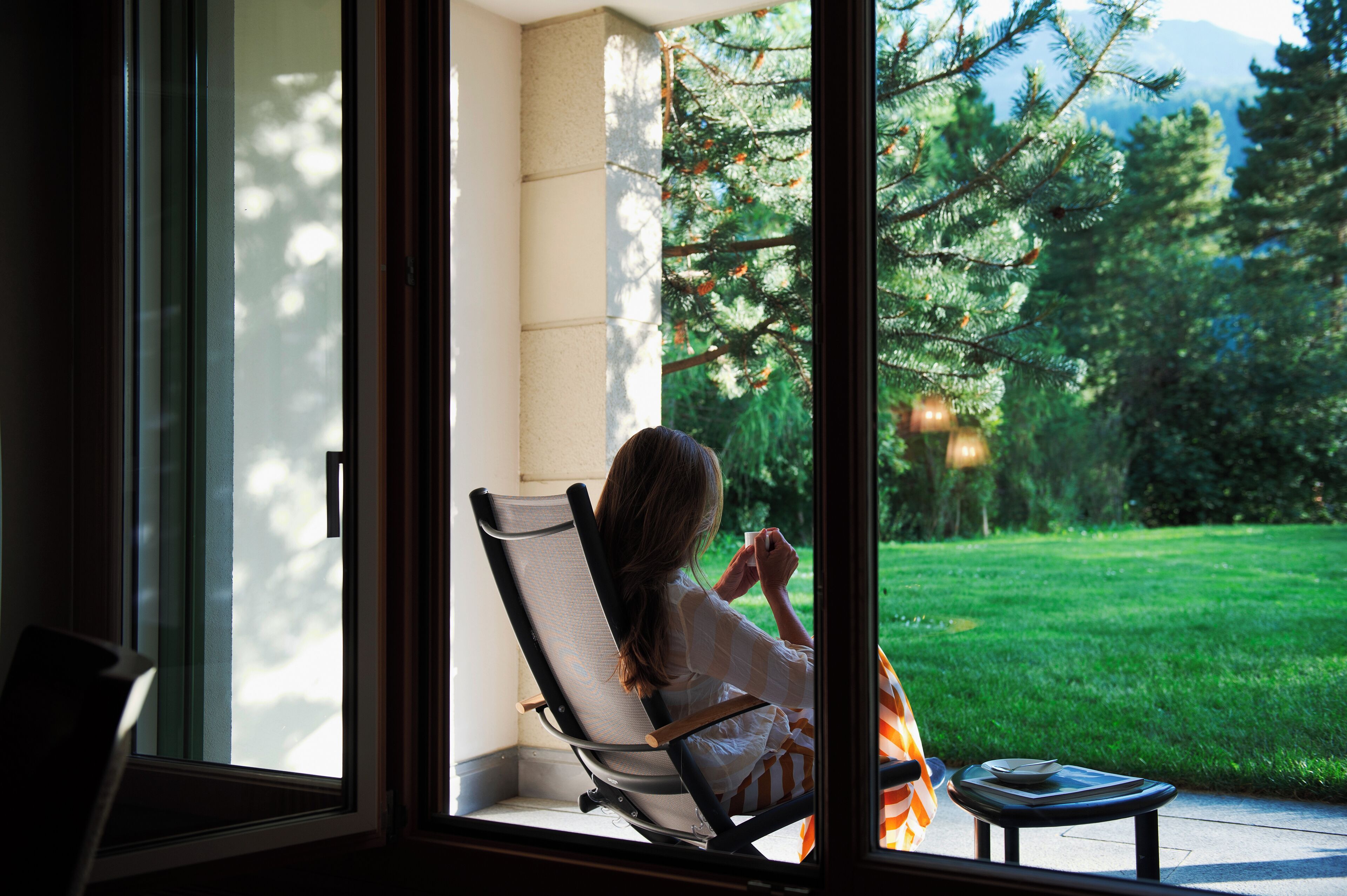 A person sitting in a chair by a window with a view of a green lawn and trees.