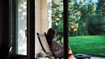 A person sitting in a chair by a window with a view of a green lawn and trees.