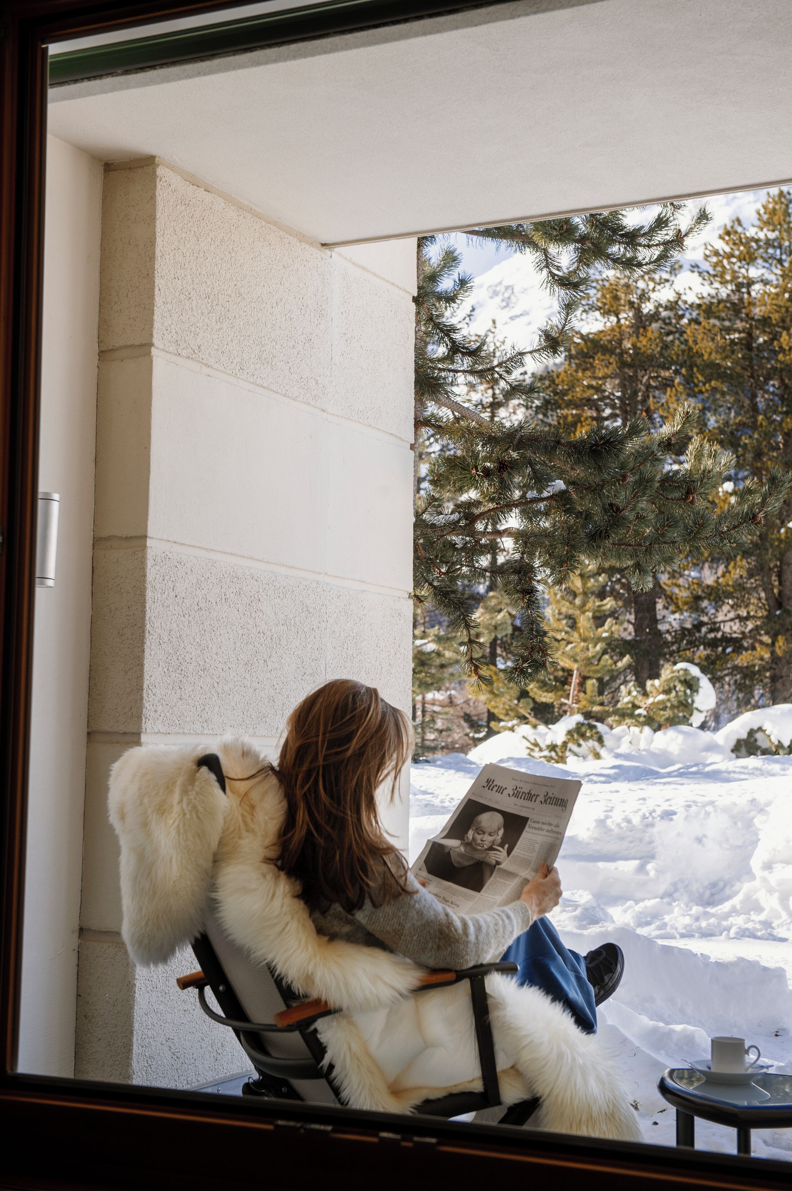 A person sitting on a chair reading a newspaper with a snowy outdoor view.