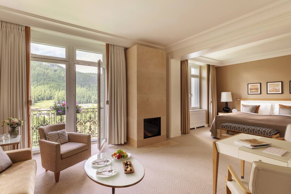A hotel room with a large window, a fireplace, a bed, a desk, and a dining table.