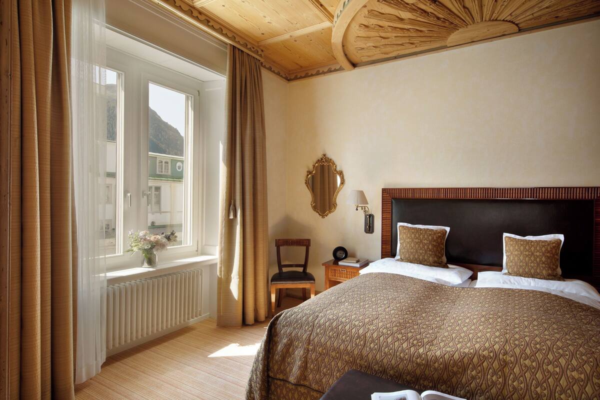 A hotel room with a large bed, a wooden headboard, a window with curtains, a chair, and a nightstand with a lamp.