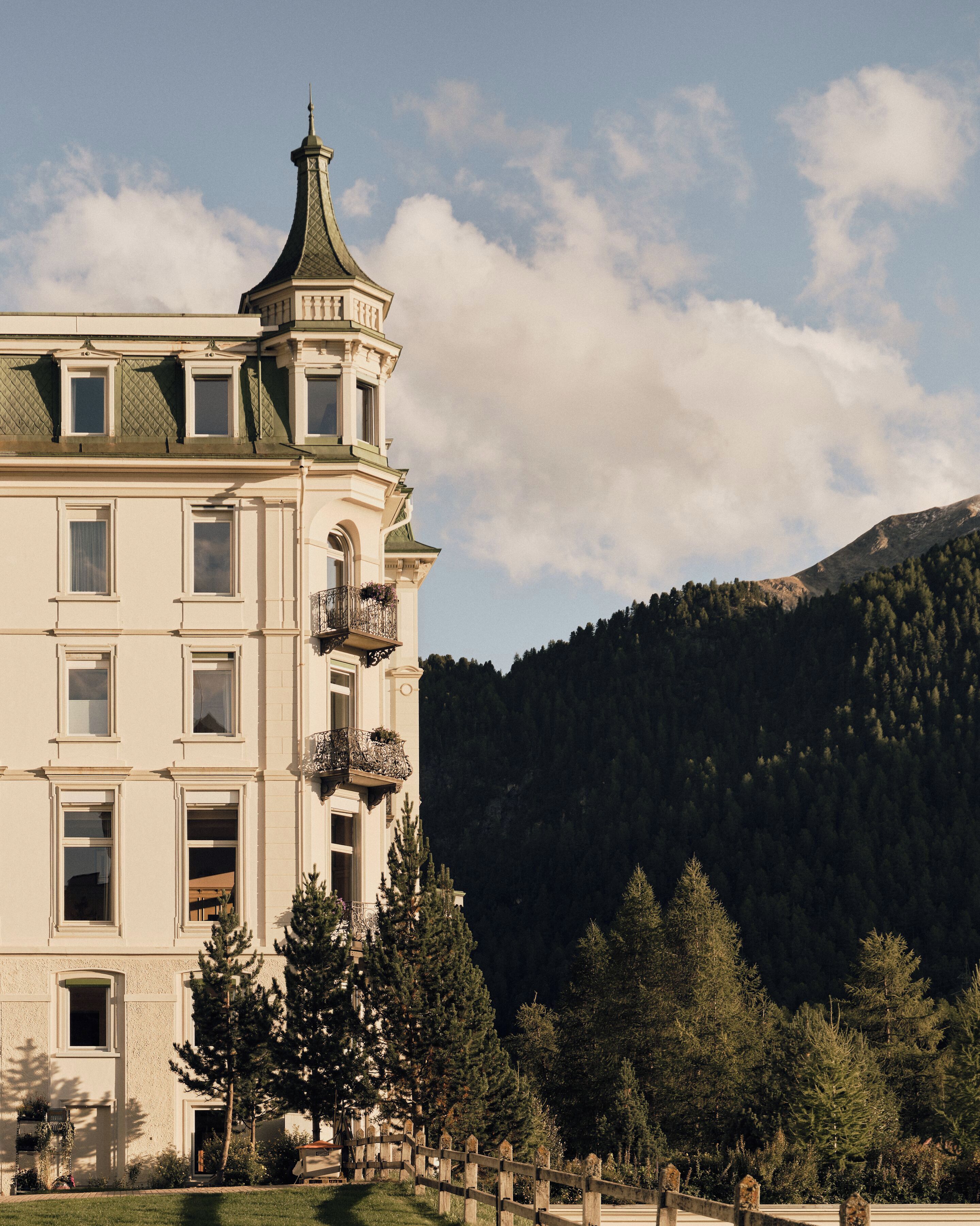 Un edificio grande de varios pisos con una torre destacada, rodeado por un paisaje montañoso y boscoso.