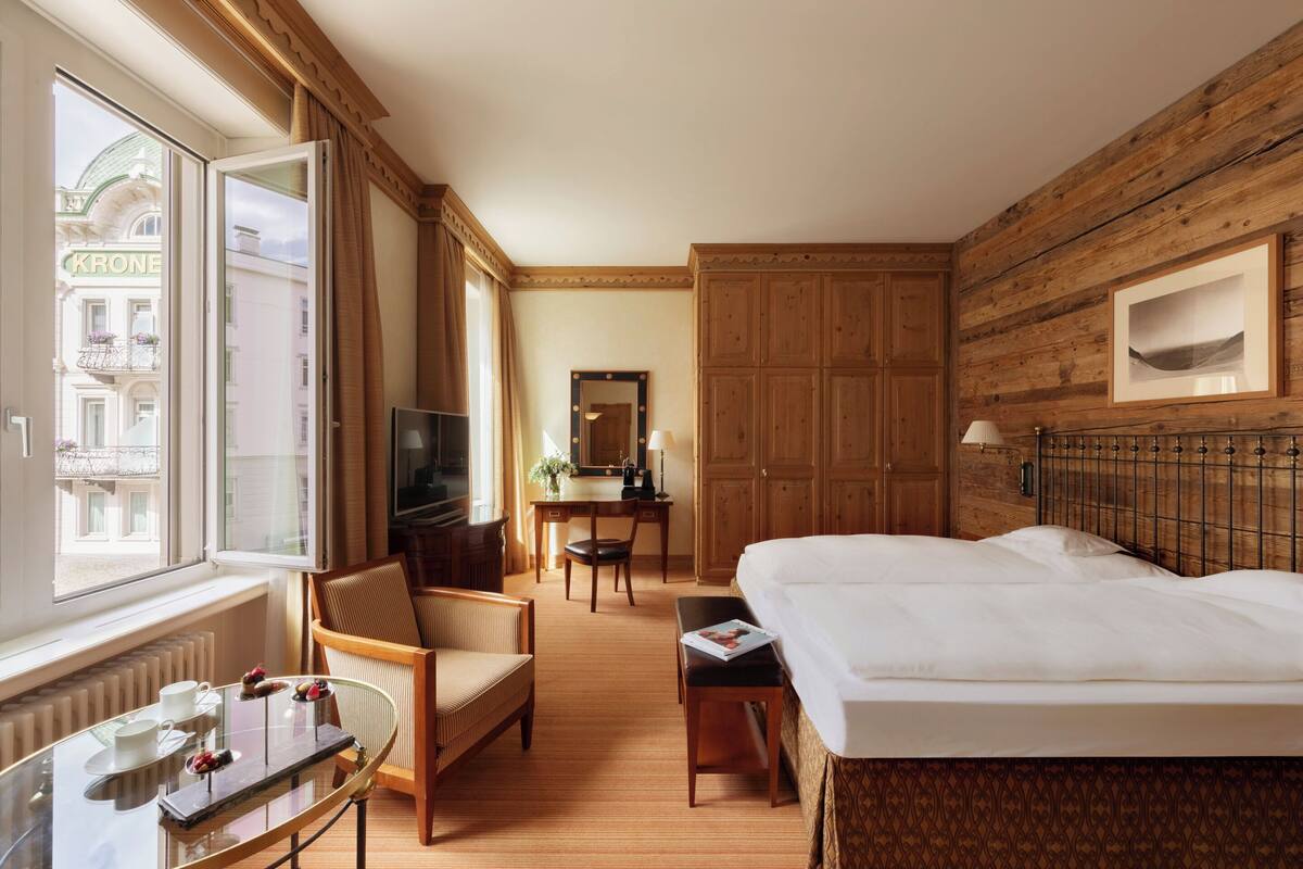 A hotel room with a bed, a desk, a chair, a television, and a window with a view of buildings.