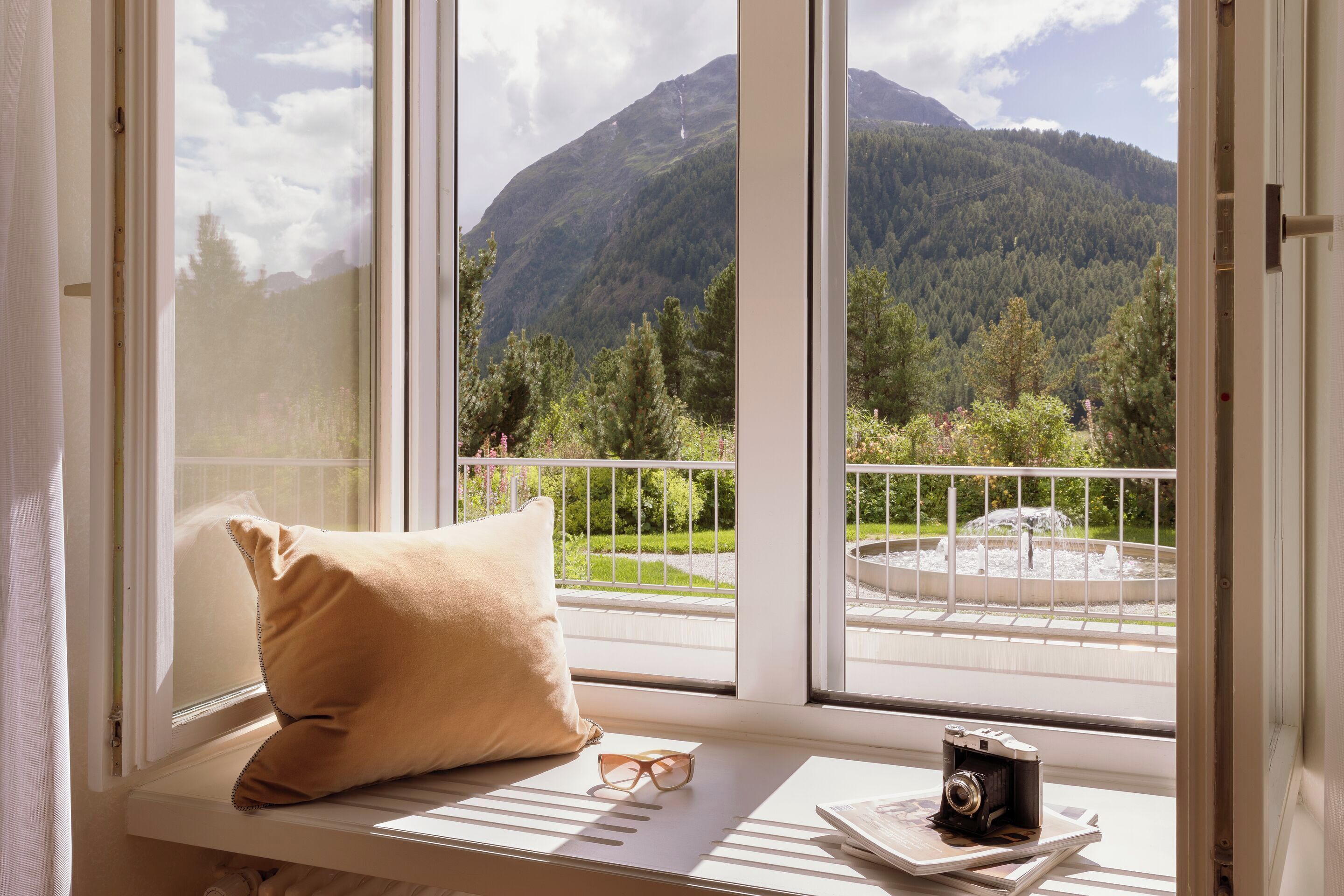 A window seat with a cushion, a pair of glasses, and a camera on a table, overlooking a mountainous landscape and a fountain.