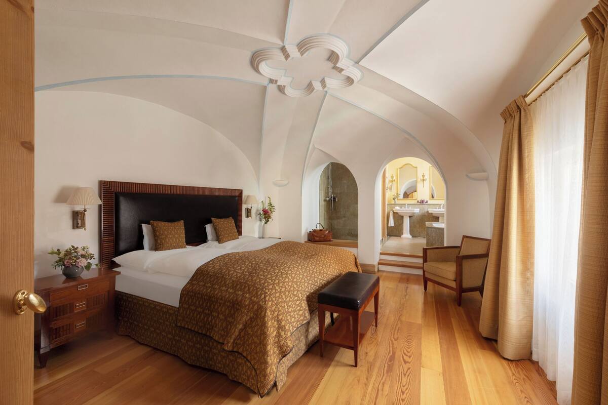 A bedroom with a large bed, wooden floor, a bench, a chair, a nightstand, and a dresser.