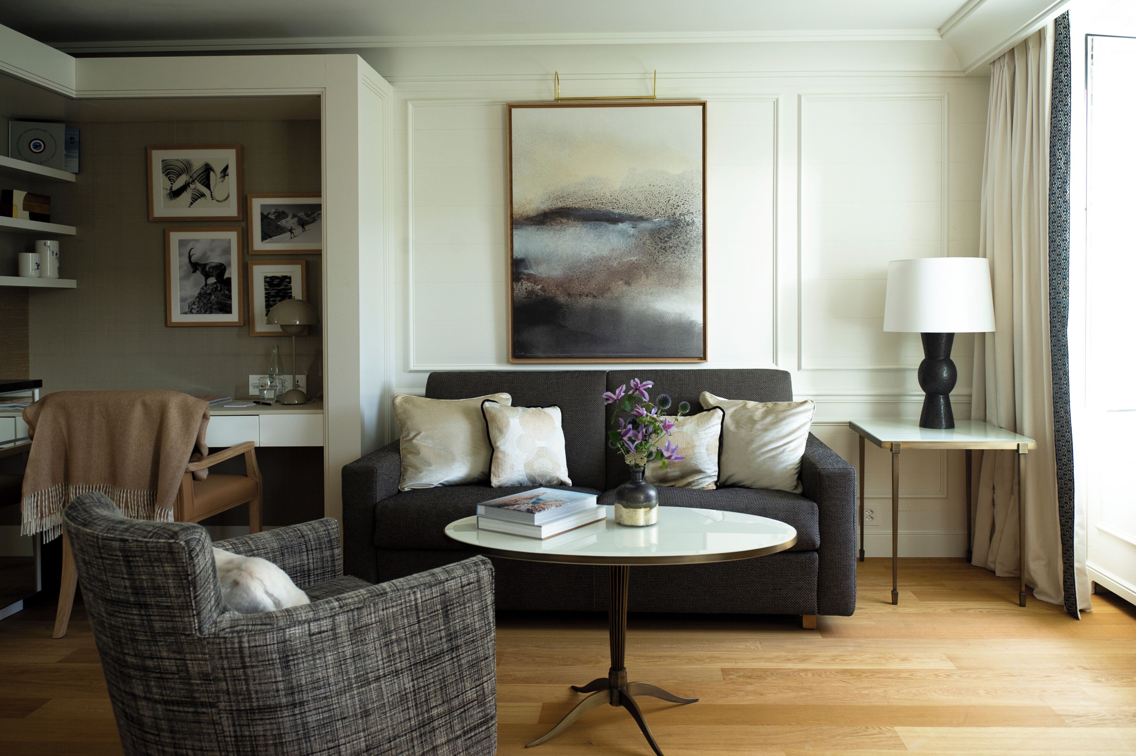 A living room with a sofa, armchair, and coffee table. There is a desk with a computer and a lamp.