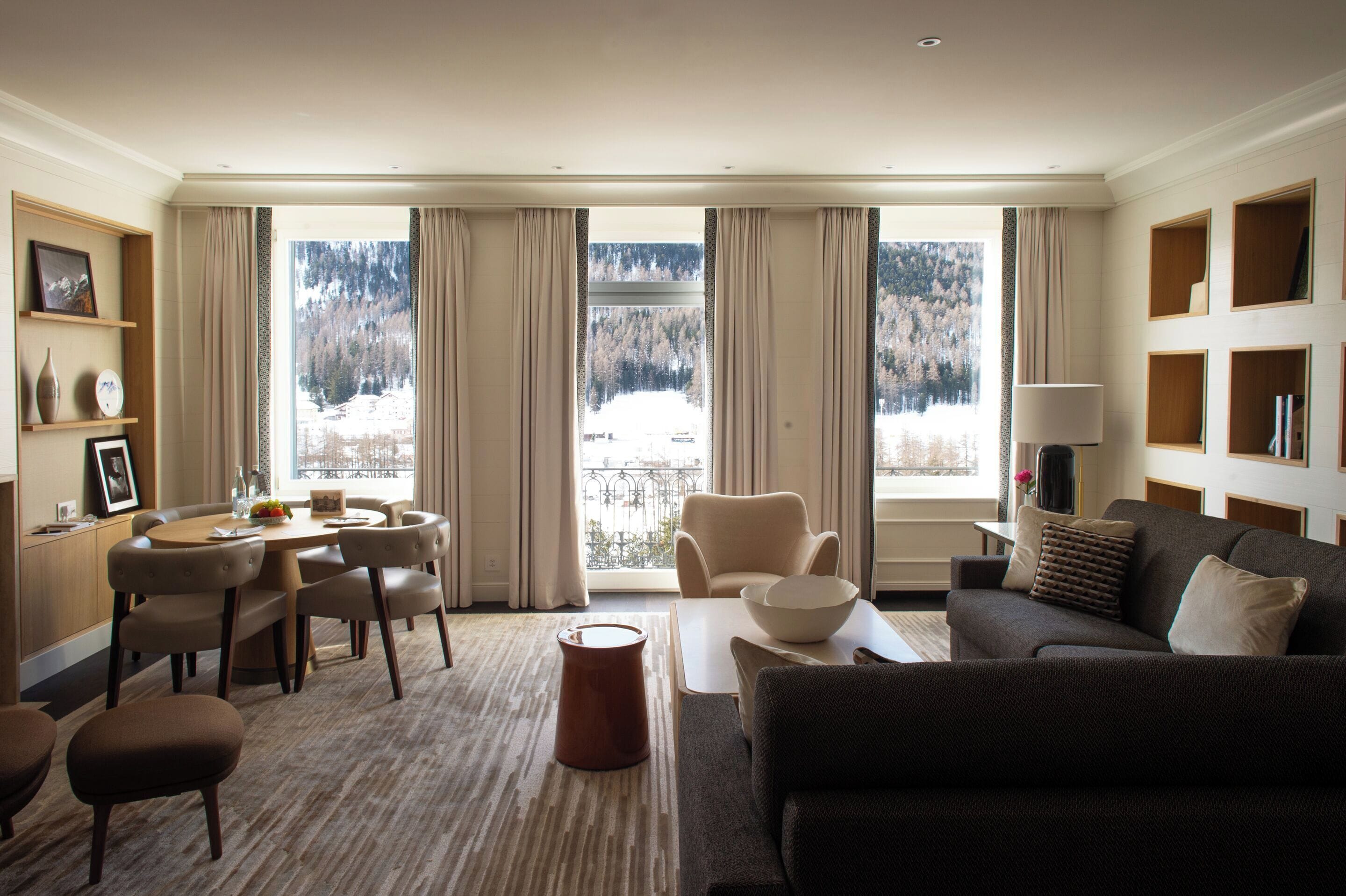 Habitación de hotel moderna con zona de comedor, sofá y vistas a un paisaje nevado a través de amplias ventanas.