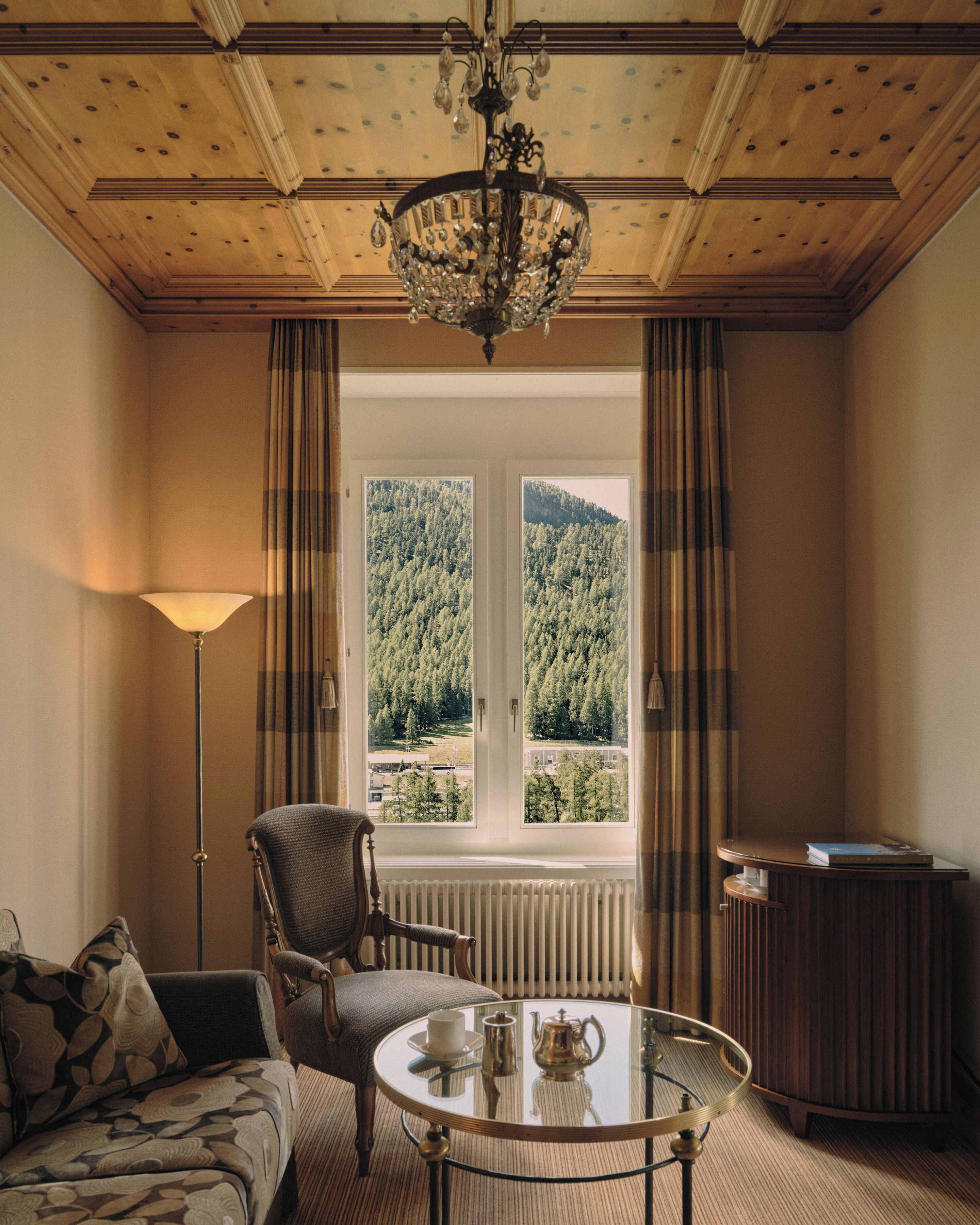 Een kamer met uitzicht op een bos, een kroonluchter, een tafel met theeservies, een stoel en een lamp.