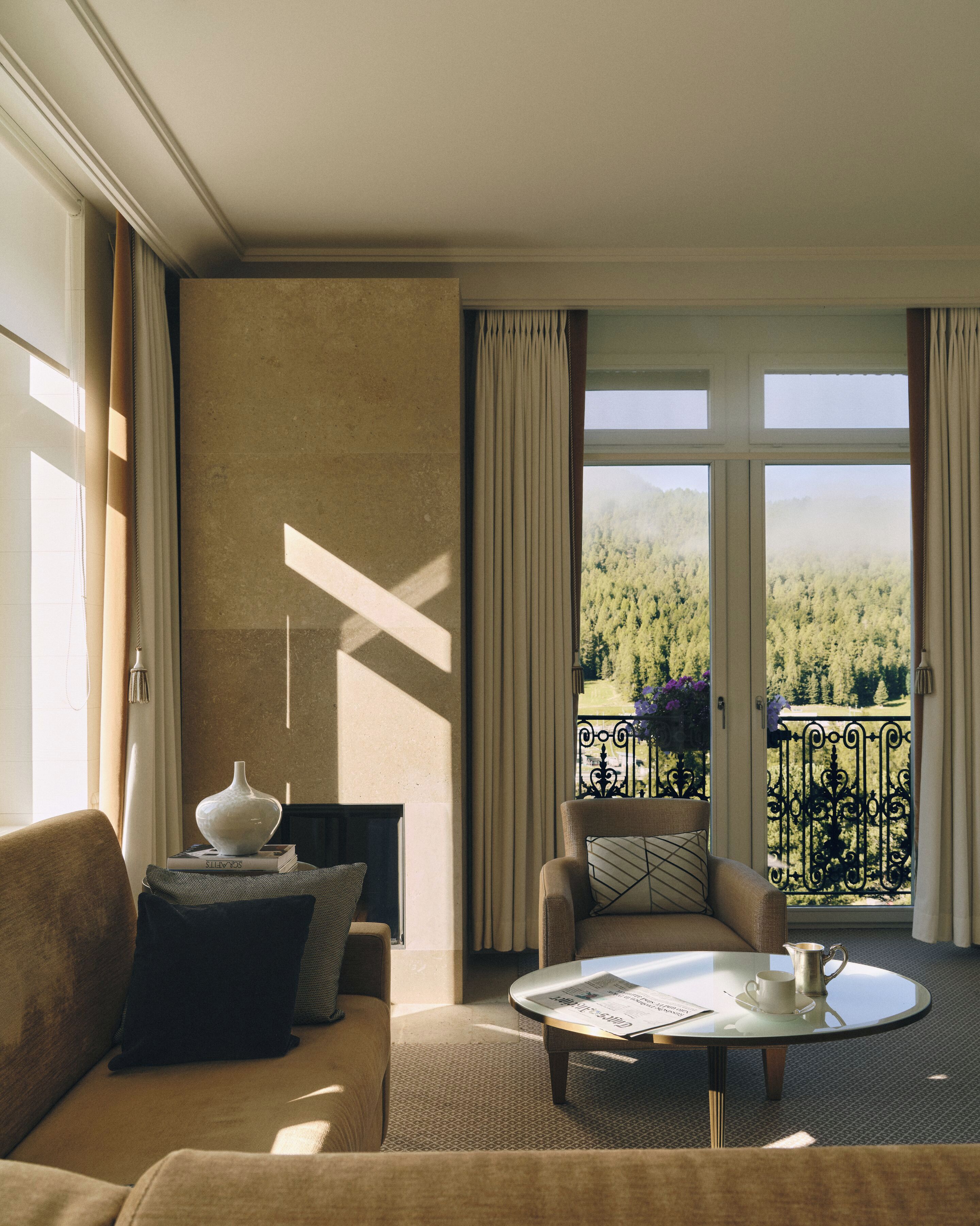 A hotel room with a balcony, a round table, a sofa, and a view of a forested landscape.