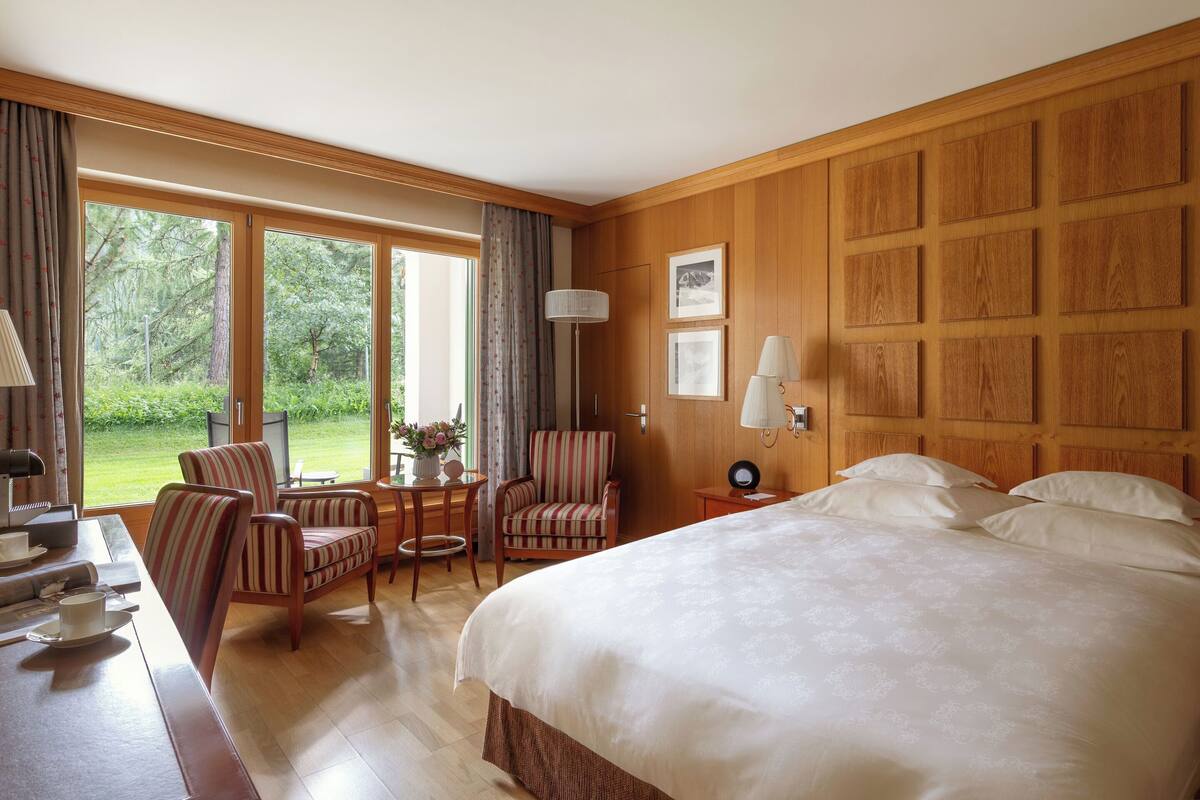 A hotel room with a large bed, two striped armchairs, a small table with a lamp, and a view of the outdoors through large windows.