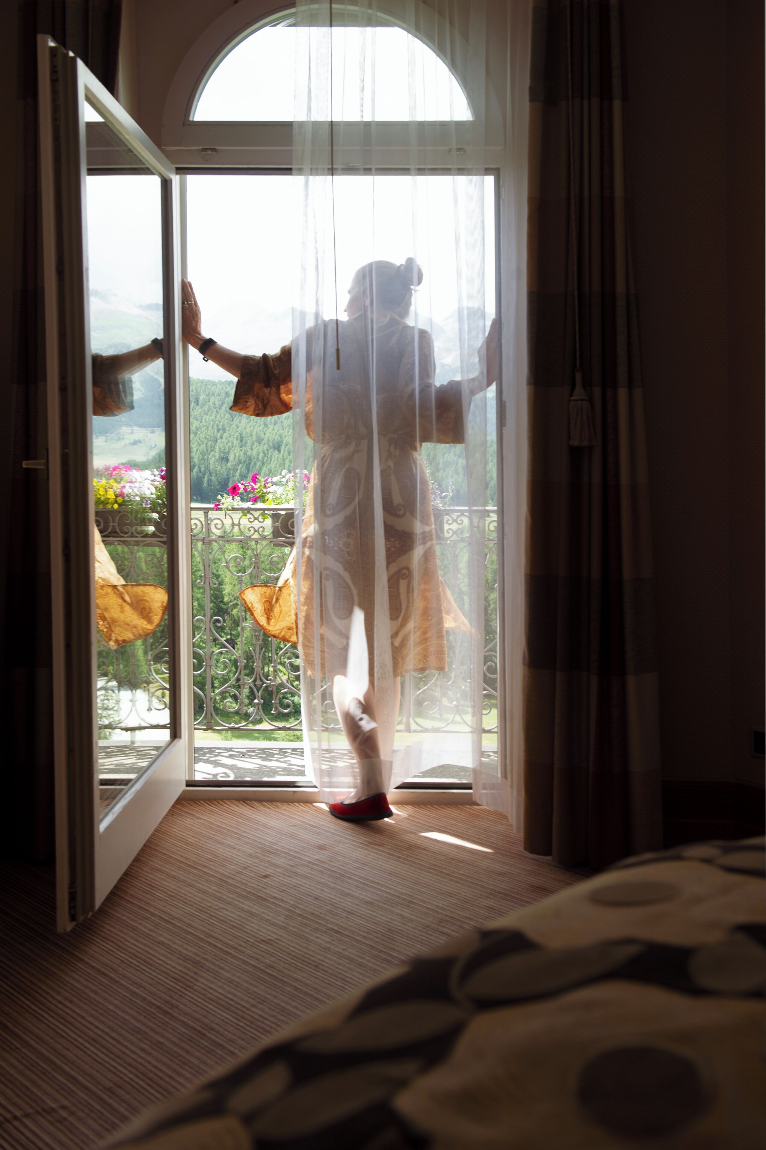 Una habitación con un ventanal amplio, una cama con colcha estampada y vistas a un balcón lleno de flores.