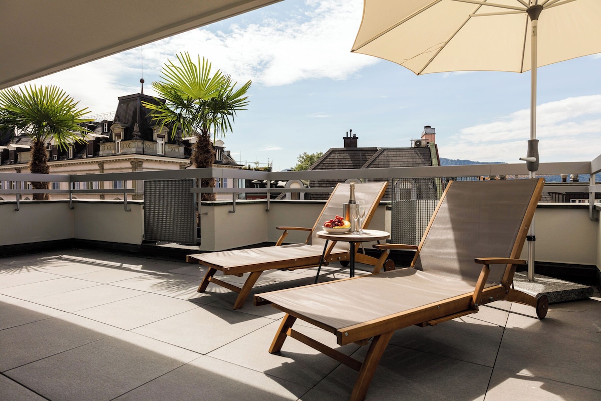 Terraço no telhado com duas cadeiras de descanso, uma mesa com bebida e frutas, e um grande guarda-sol.