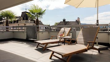 A rooftop terrace with two lounge chairs, a table with a drink and fruit, and a large umbrella.