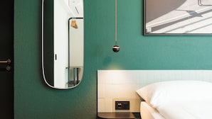 Une chambre moderne avec un mur vert, un grand miroir, un lit, une table de chevet et un luminaire fixé au mur.