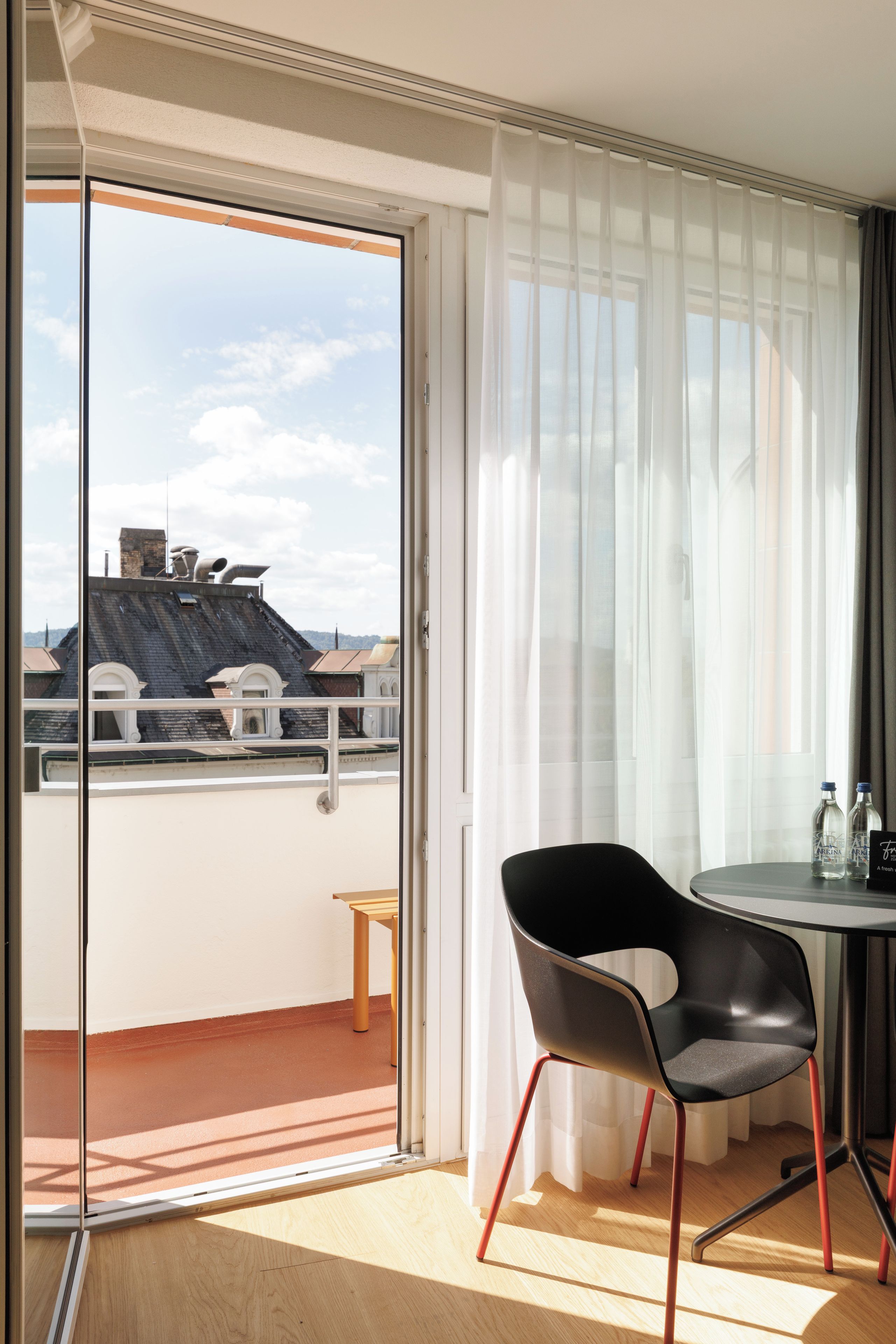 Una habitación con puerta corrediza de vidrio que da a un balcón, una mesa redonda con dos sillas y un taburete pequeño.