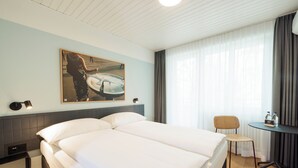 A hotel room with a large bed, a small table with a chair, a painting on the wall, and a window with curtains.