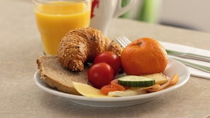 A breakfast plate with bread, a croissant, an orange, tomatoes, and a glass of orange juice on a table.