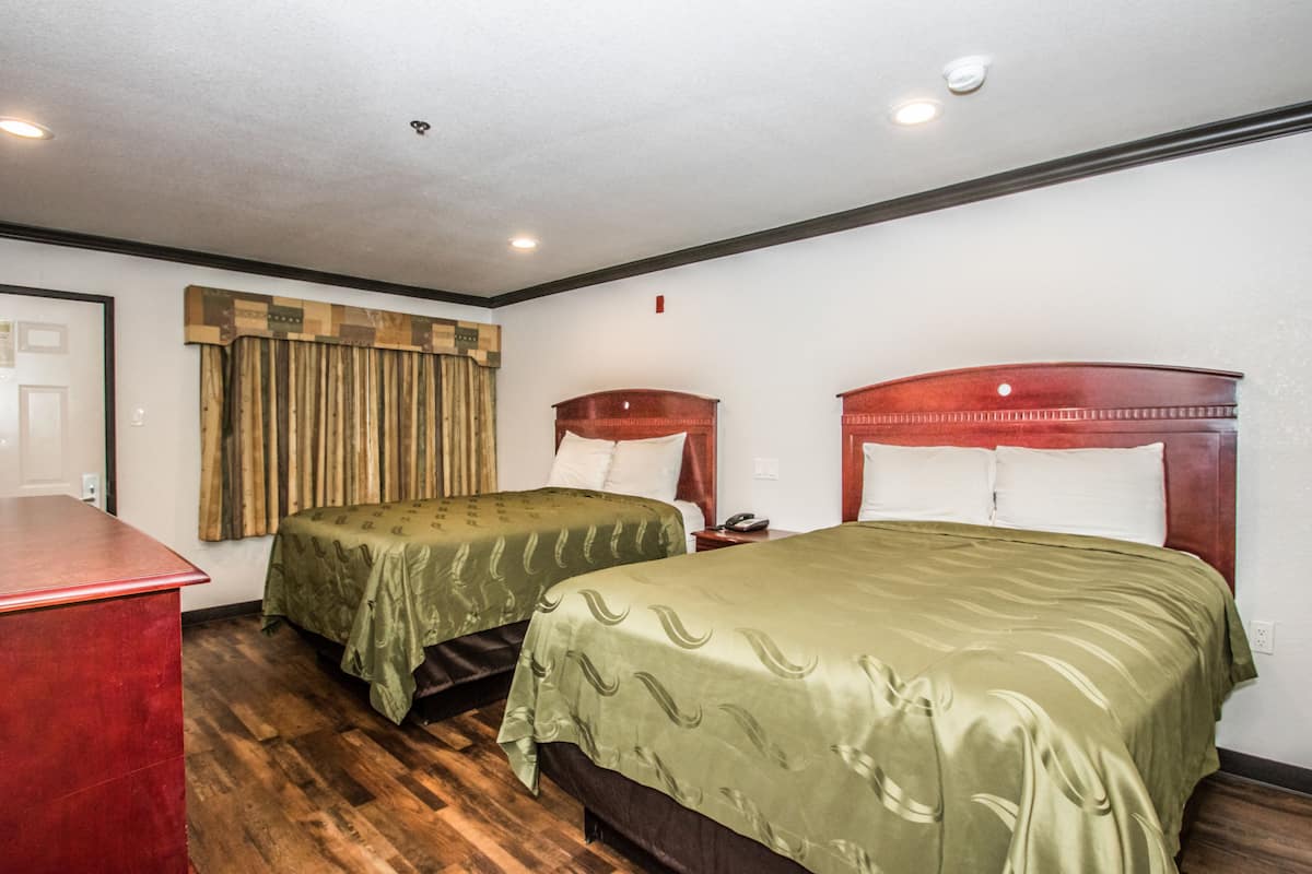 A hotel room with two beds, wooden flooring, a desk, and a window with curtains.