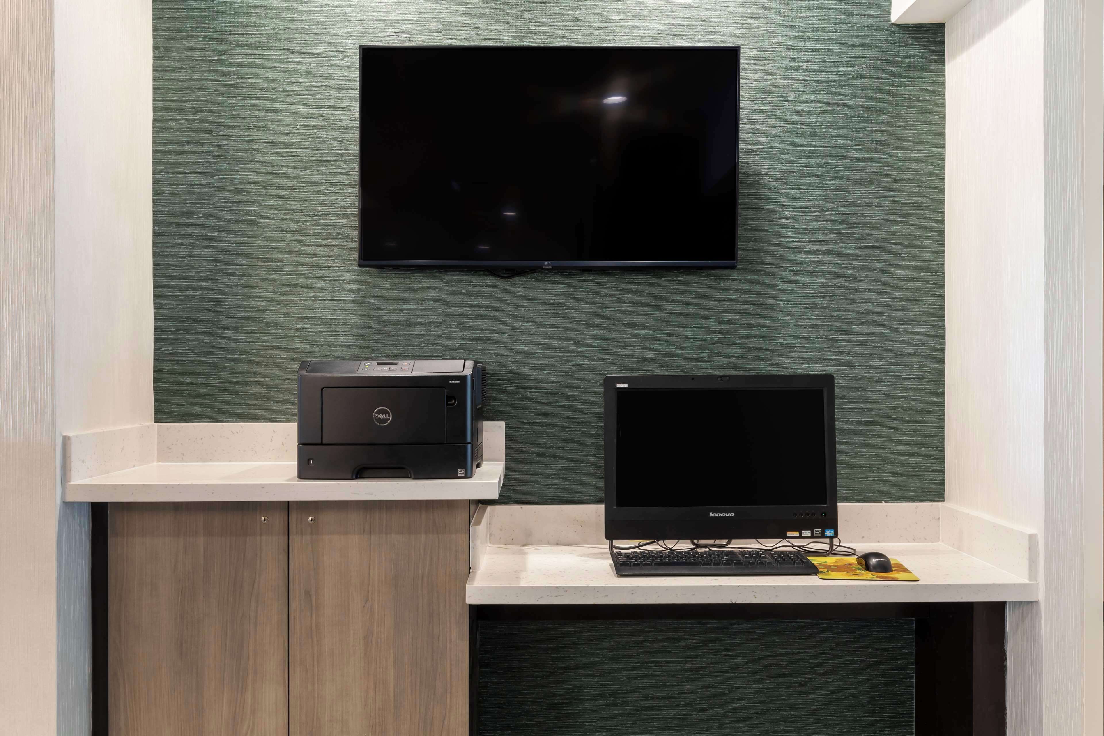 A workspace with a flat-screen TV mounted on the wall, a Dell printer, and a Lenovo laptop on a white desk.