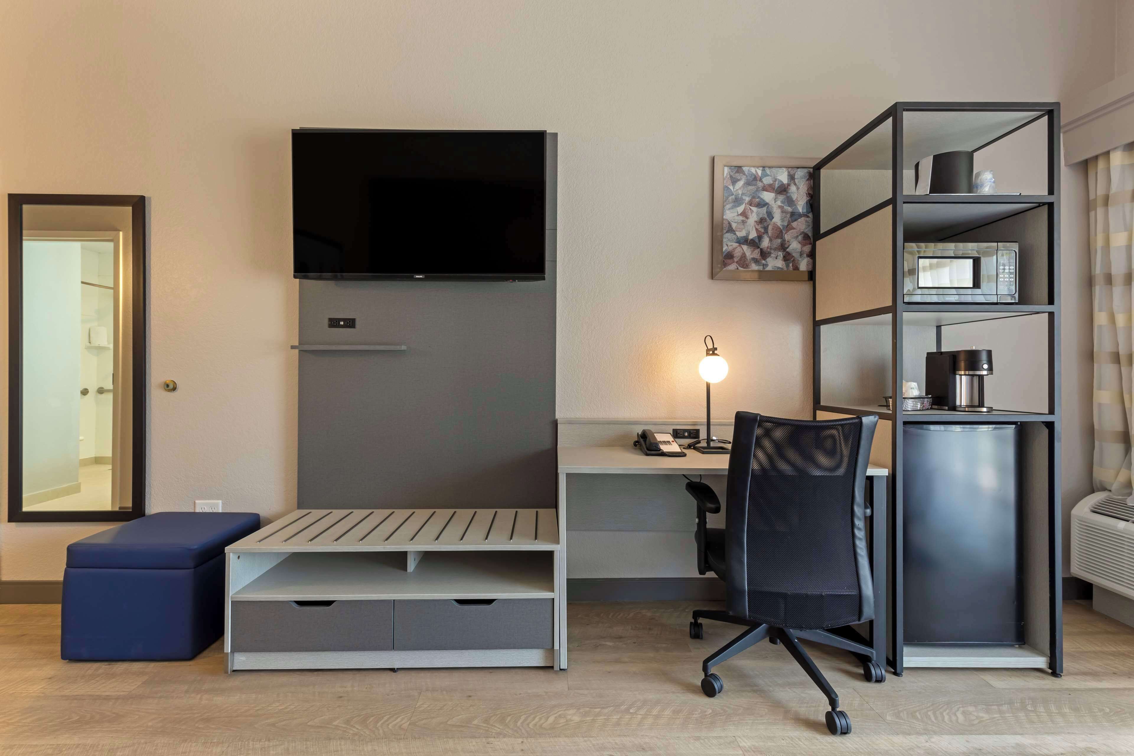 Una habitación de hotel moderna con un televisor de pantalla plana montado en la pared, un escritorio con una silla, un mueble bajo y una mesita de noche con lámpara.