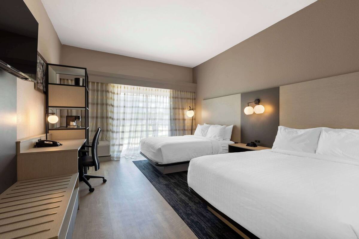 Une chambre d’hôtel avec deux lits, un bureau, une chaise, un miroir et une fenêtre avec des rideaux.