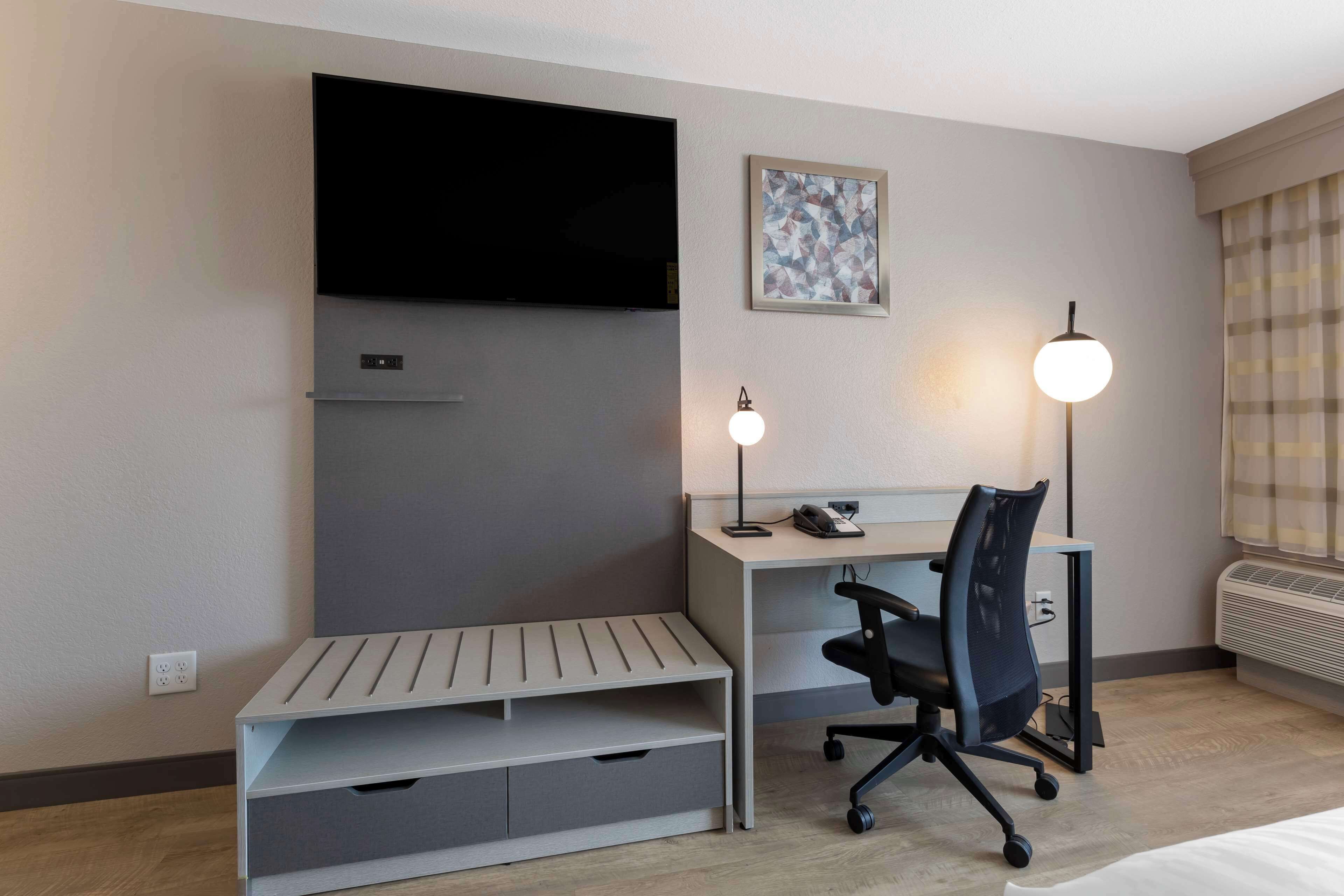 Habitación de hotel con cama, escritorio y silla, televisor en la pared, un pequeño banco, lámpara de escritorio y un cuadro enmarcado.