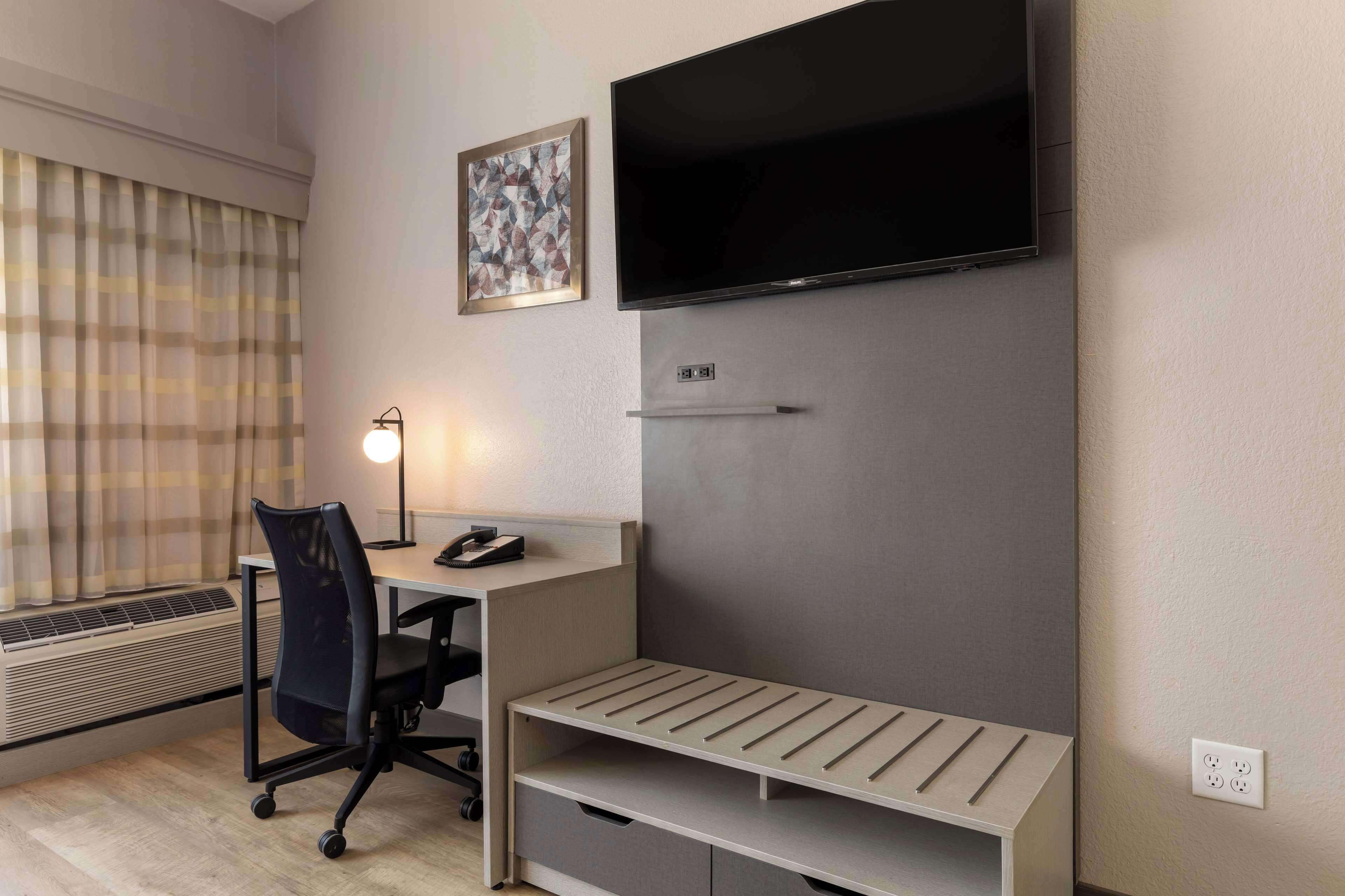 A hotel room with a desk, chair, TV, and a small shelf.