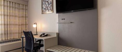 A hotel room with a desk, chair, TV, and a small shelf.