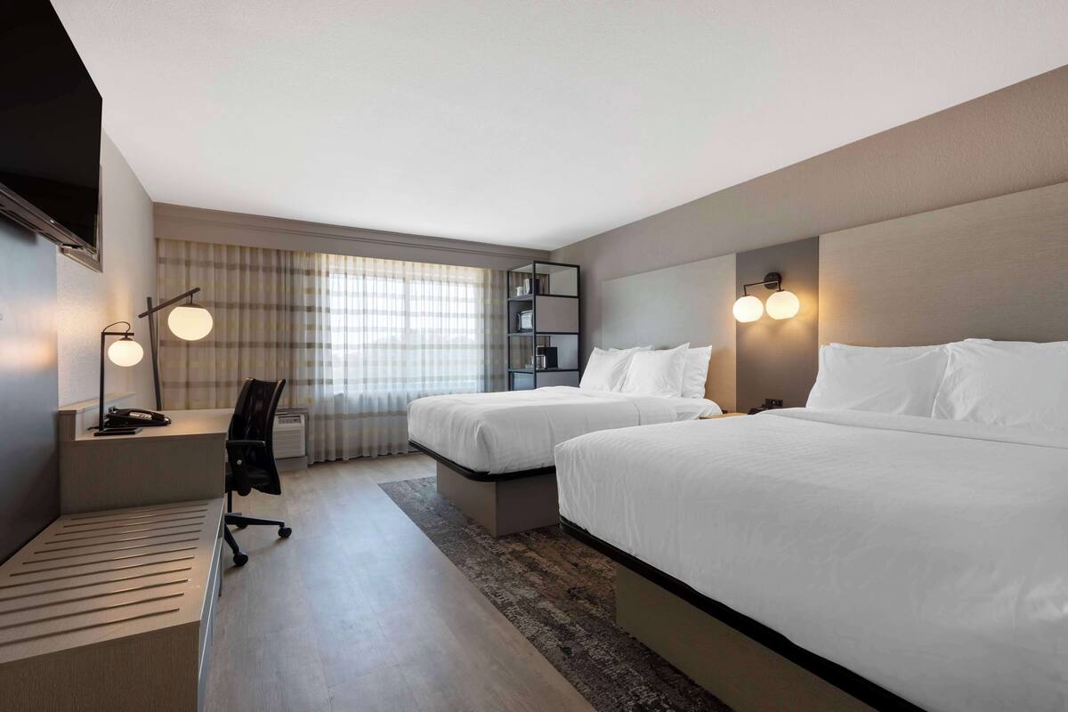 Une chambre d’hôtel avec deux lits, un bureau, une chaise, une bibliothèque et une grande fenêtre avec des rideaux.