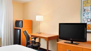 Una habitación de hotel con una cama, un escritorio con monitor de computadora, una silla, una lámpara de escritorio, un televisor, un refrigerador y una obra de arte en la pared.