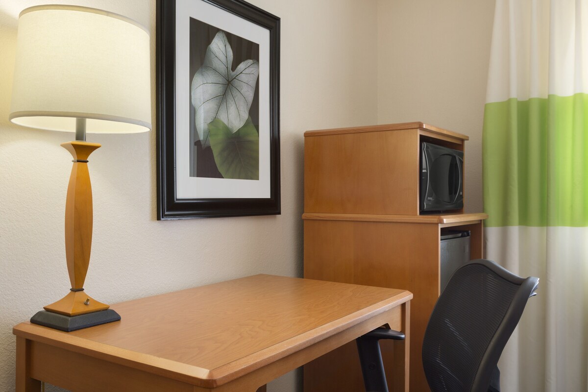 A hotel room with a wooden desk, a black office chair, a framed picture of a flower, a lamp, and a microwave.