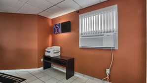 A room with orange walls, a tiled floor, a black desk with printers, a mounted air conditioner, and a window with blinds. - Motel 6 Windsor, ON (Windsor)