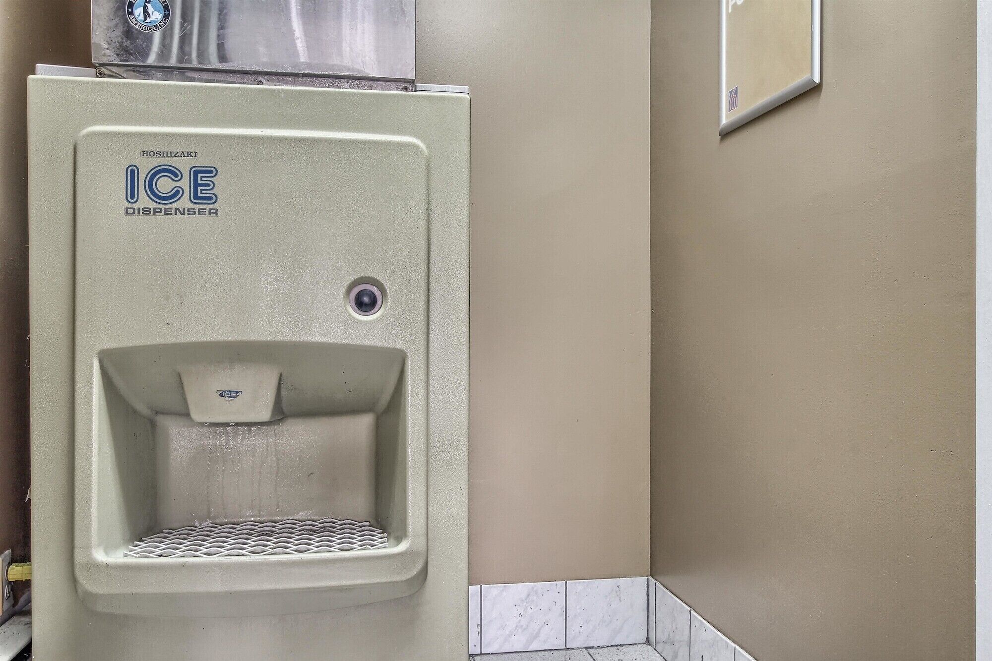 A Hoshizaki ice dispenser in a restroom with a beige wall and a tiled floor.