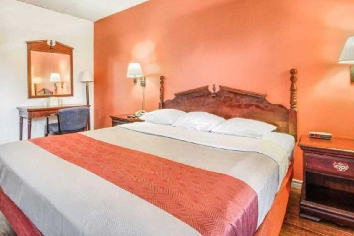 A hotel room with a large bed, a wooden headboard, a desk with a chair, two bedside lamps, and a mirror on the wall.