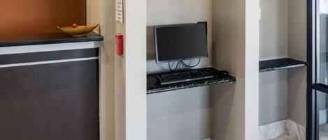 Zona de recepción de un hotel con un mostrador oscuro, un televisor de pantalla plana montado en la pared y un estante pequeño con un teclado.