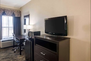 Een hotelkamer met een flatscreen televisie aan de muur, een bureau met een stoel, een lamp en een groot raam met gordijnen.