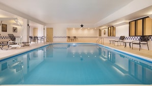 En stor indendørs swimmingpool med fliserede vægge og et skakmønster. Der er liggestole og borde ved poolen.