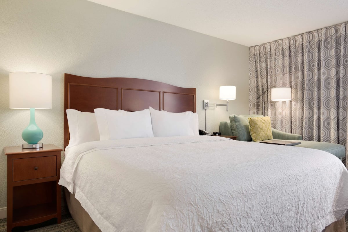 A hotel room with a large bed, a nightstand with a lamp, a chair, and patterned curtains.