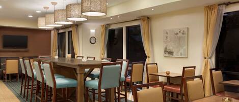 A hotel dining area with multiple tables and chairs, pendant lighting, a clock, and a large window with curtains.