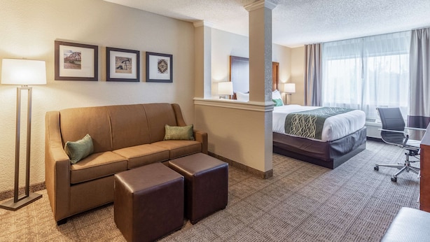A hotel room with a sofa, ottoman, bed, desk, and lamp. - Comfort Inn South Tulsa - Woodland Hills (Tulsa)