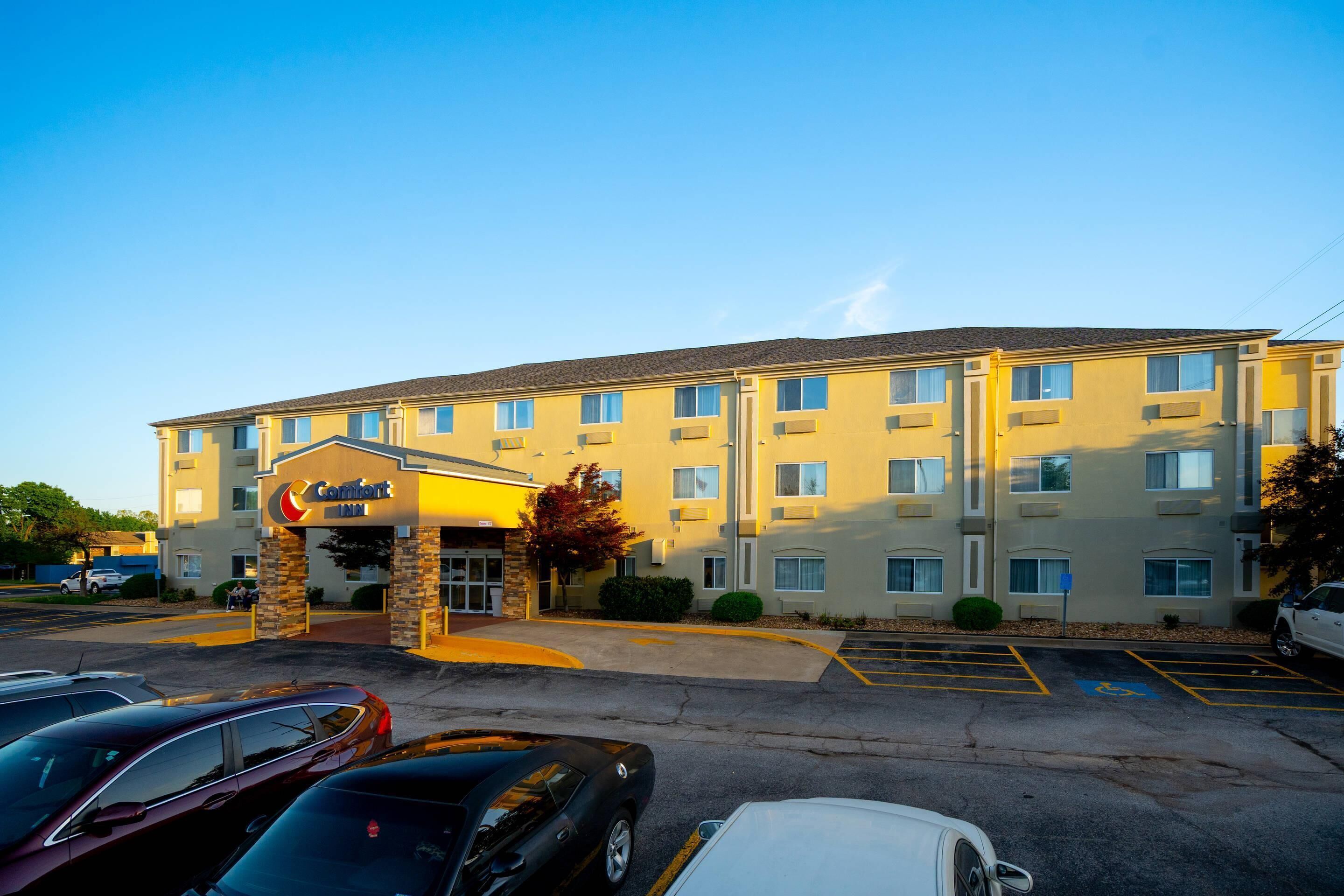 Un hotel de varios pisos con techo plano, una entrada central con arco de piedra y un estacionamiento con varios coches.