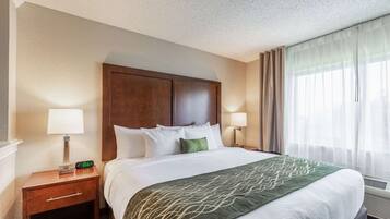 Quarto de hotel com uma cama grande, duas mesas de cabeceira, um criado-mudo, um cobertor verde e uma janela com cortinas translúcidas.