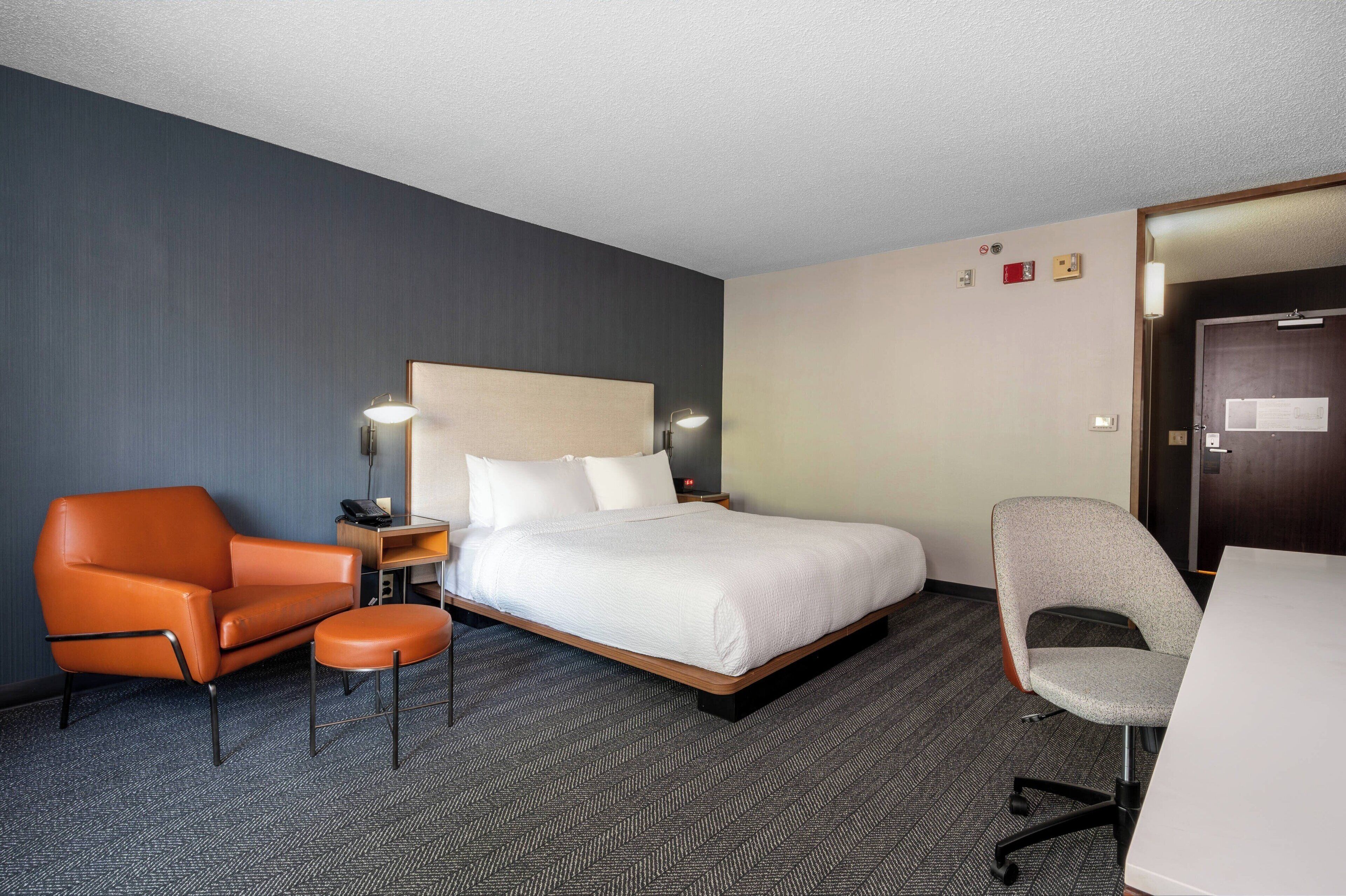 Quarto de hotel com cama, duas cadeiras, escrivaninha e piso coberto de carpete.