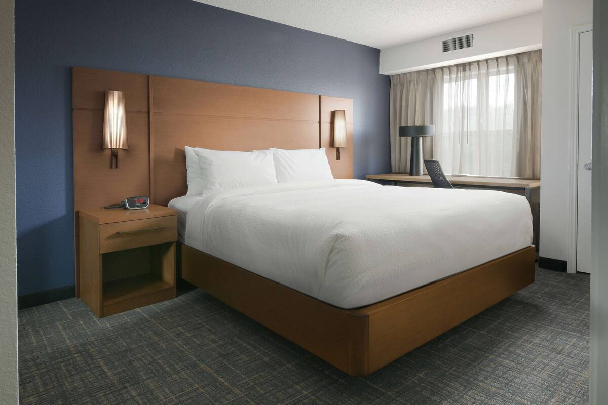 Ein Hotelzimmer mit einem großen Bett, einem Nachttisch mit Lampe, einem Schreibtisch mit Stuhl und einem Fenster mit Vorhängen.