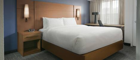 Habitación de hotel con una cama grande, una mesita de noche con lámpara, un escritorio con silla y una ventana con cortinas.