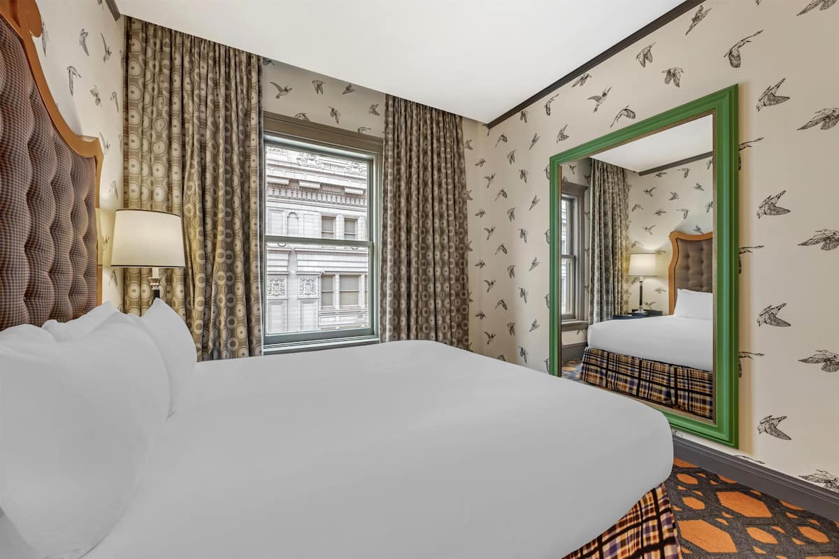 A hotel room with a large bed, a mirror, a window with curtains, and a view of a building outside.