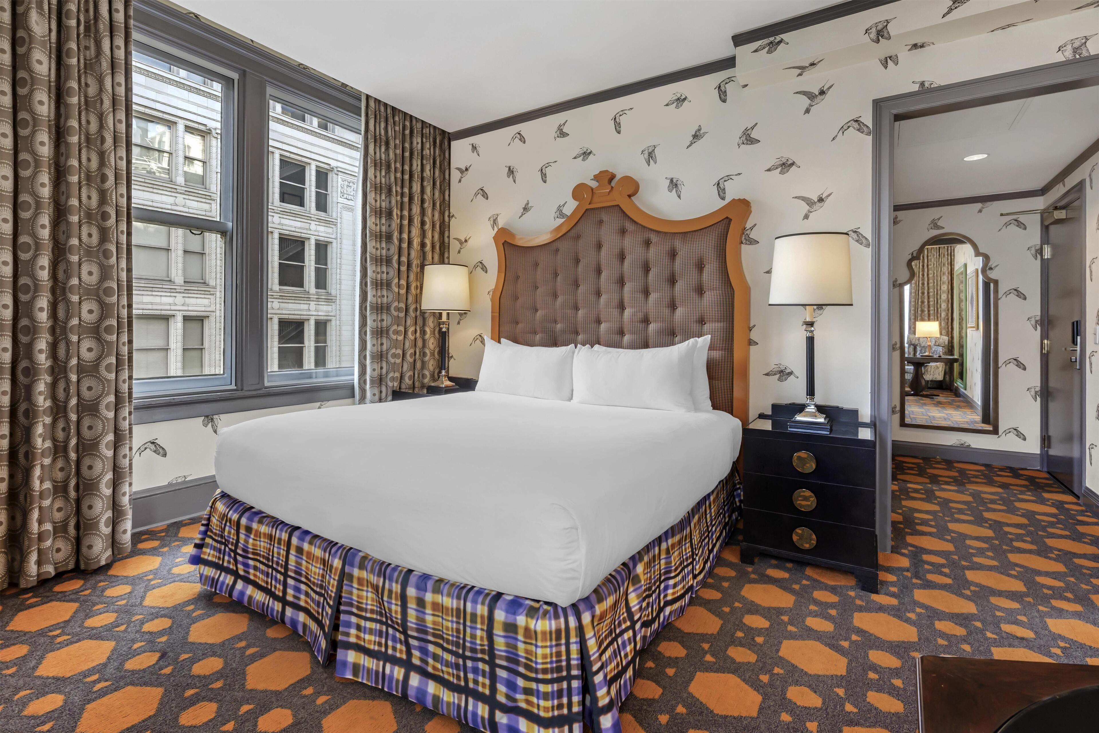 A hotel room with a large bed, two bedside lamps, a dresser, a mirror, and patterned wallpaper.